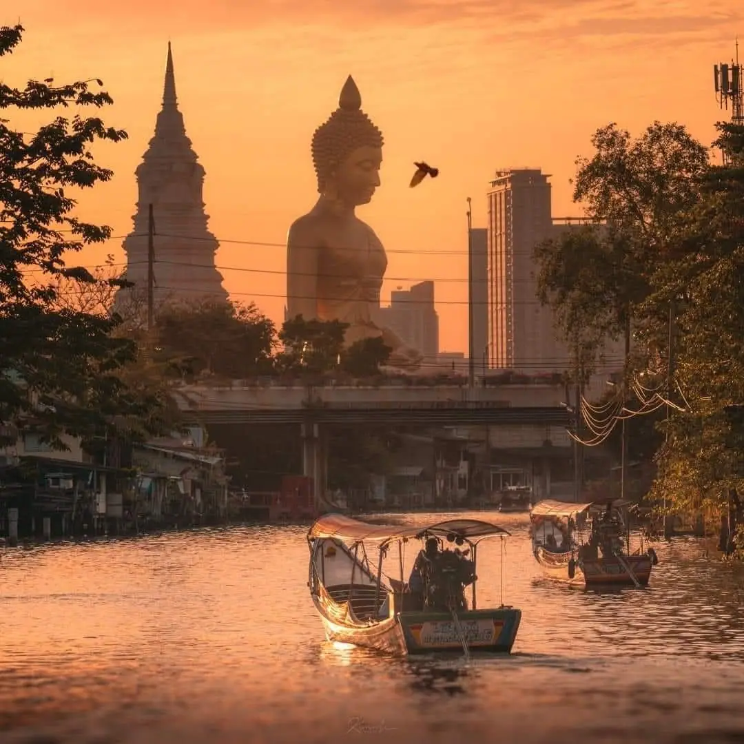 คลองยามพระอาทิตย์ตก มีเรือหางยาว และพระพุทธรูปองค์ใหญ่อยู่หลังแนวตึกของเมือง