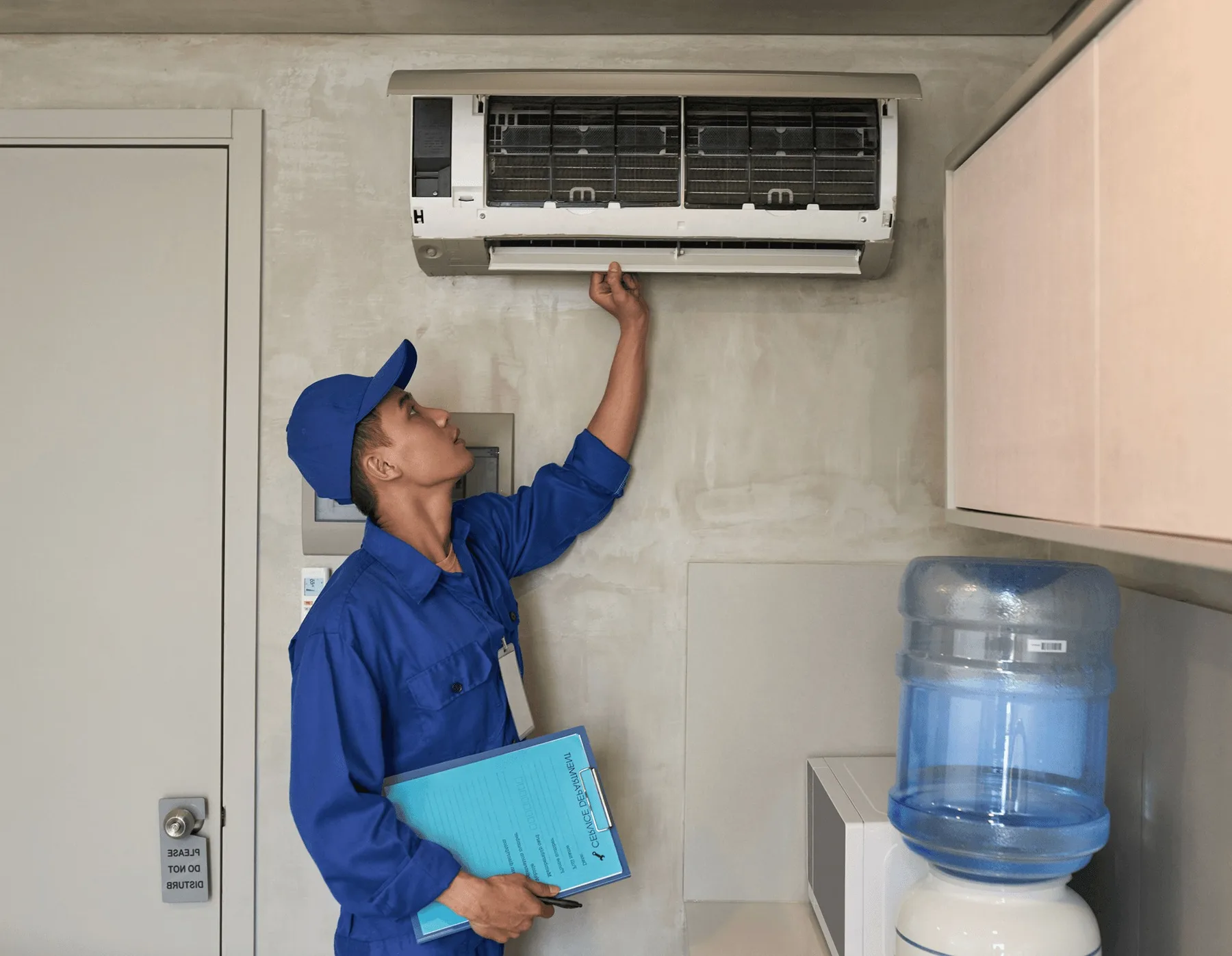 A technician in blue reaching up to a wall unit indoors.
