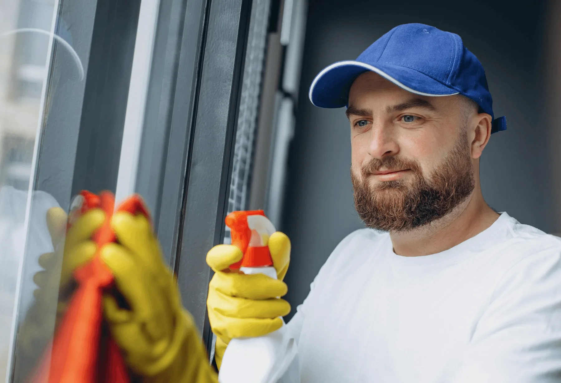 A bearded cleaner in a blue cap spraying a window.