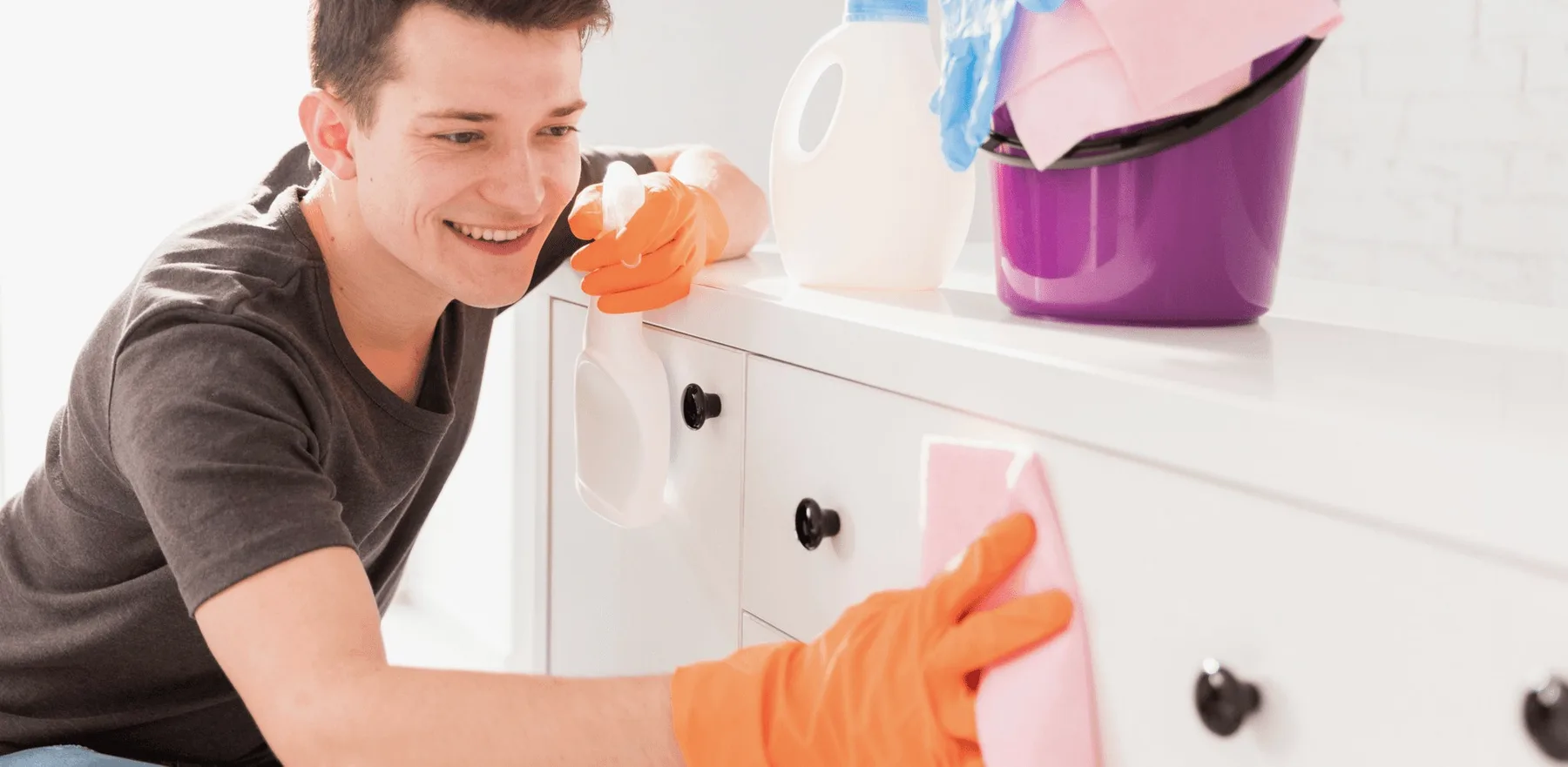 A smiling cleaner in orange gloves wiping a white cabinet.