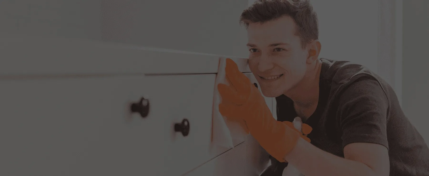 A smiling cleaner in orange gloves wiping a cabinet, under a dark veil.