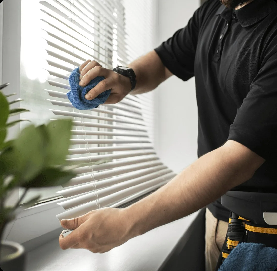 A cleaner in black wiping window blinds with a blue cloth.