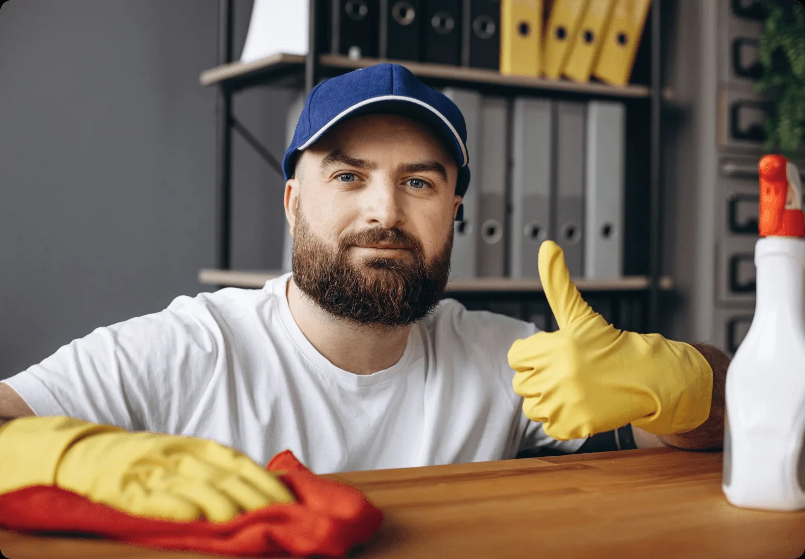 A bearded cleaner in a blue cap giving a thumbs up at a desk.