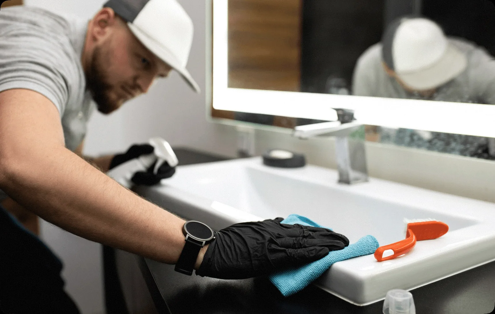 A cleaner in a white cap wiping a bathroom sink.