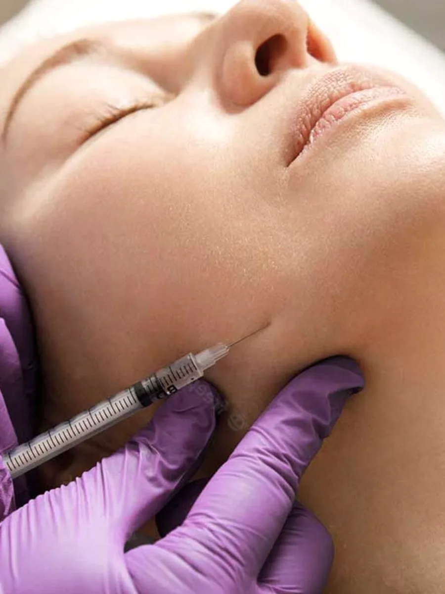 A face under a syringe held by a purple-gloved hand.