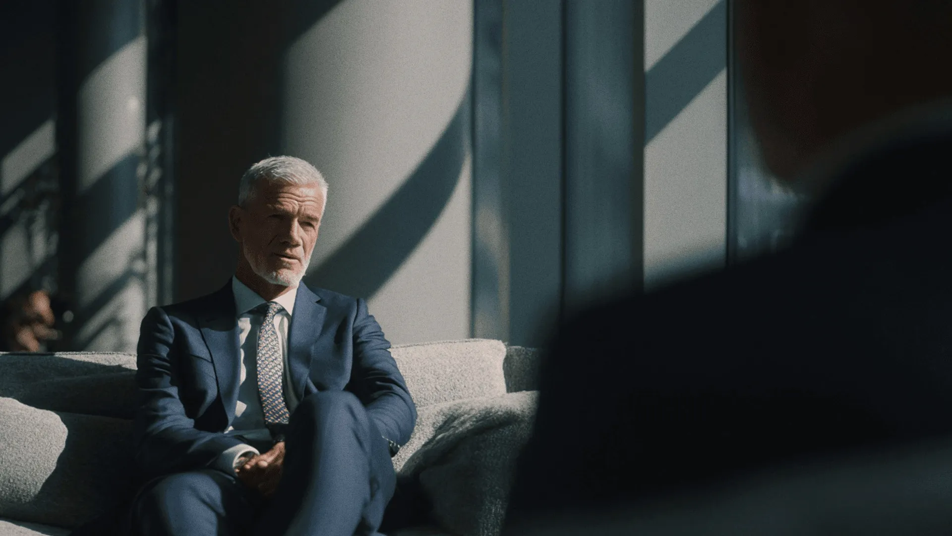 A man in a navy suit on a grey sofa in striped light.