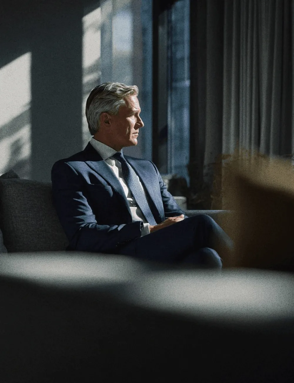 A man in a navy suit seated by a window.