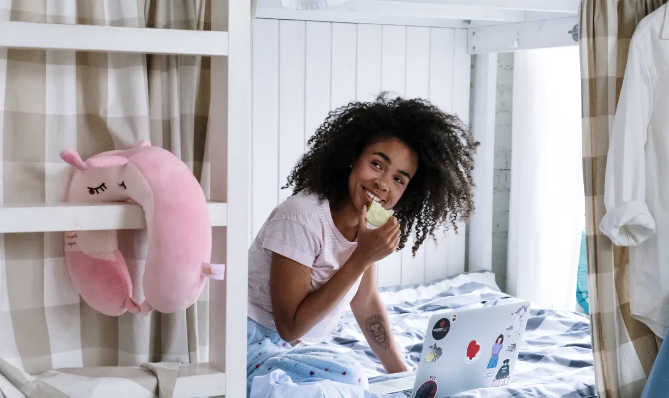 A guest eating an apple on a bunk bed beside her laptop.