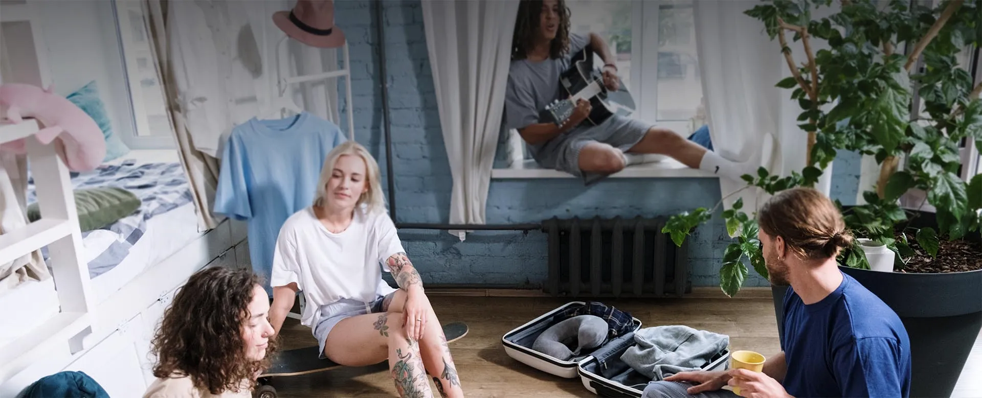Young travellers relaxing in a bright hostel room, one playing guitar on the window sill.