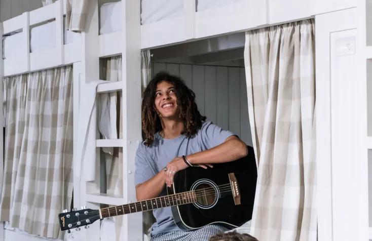 A man with a guitar smiling in a curtained bunk bed.