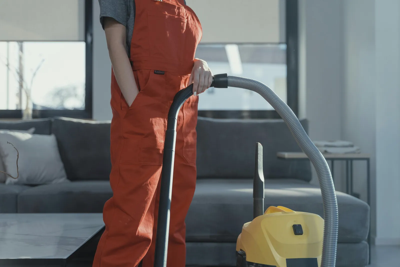 A cleaner in red overalls vacuuming beside a grey sofa.