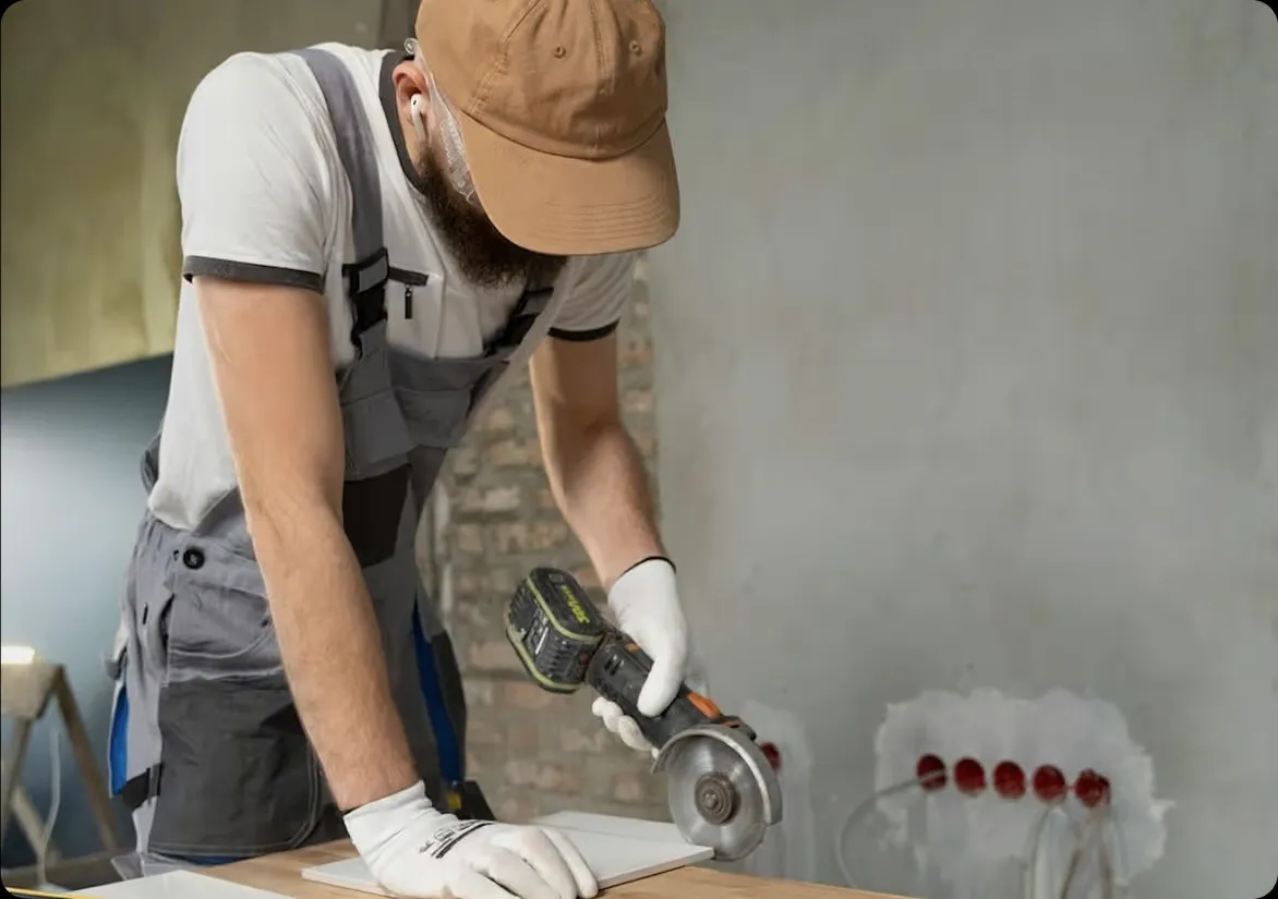 A worker in a cap and grey overalls cutting tile with a grinder.