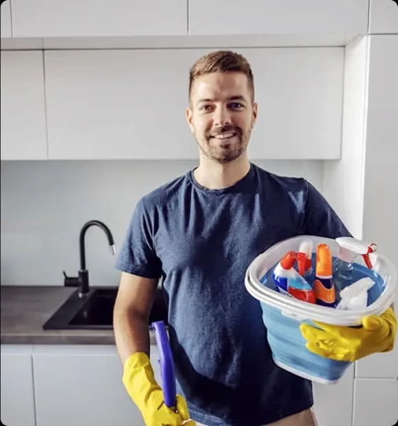 A smiling man in a navy shirt holding a basket of cleaning supplies.