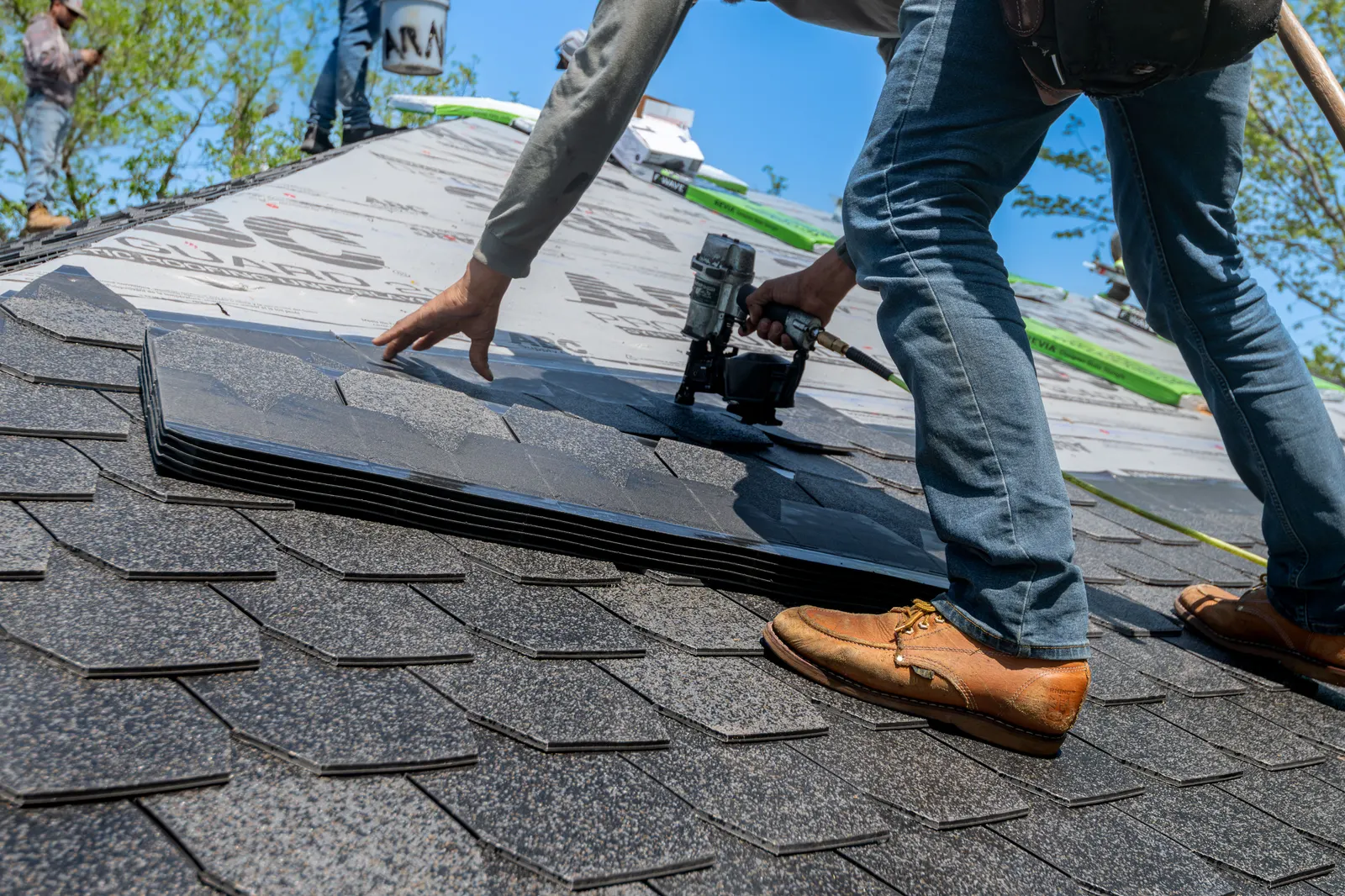 Hands laying a new shingle on a roof deck, a worker's boots in frame beside it.