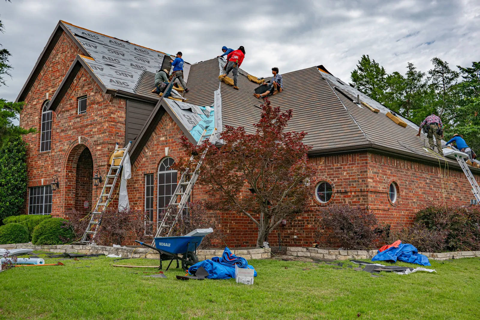 A crew re-roofing a brick house, ladders up and materials out on the lawn.