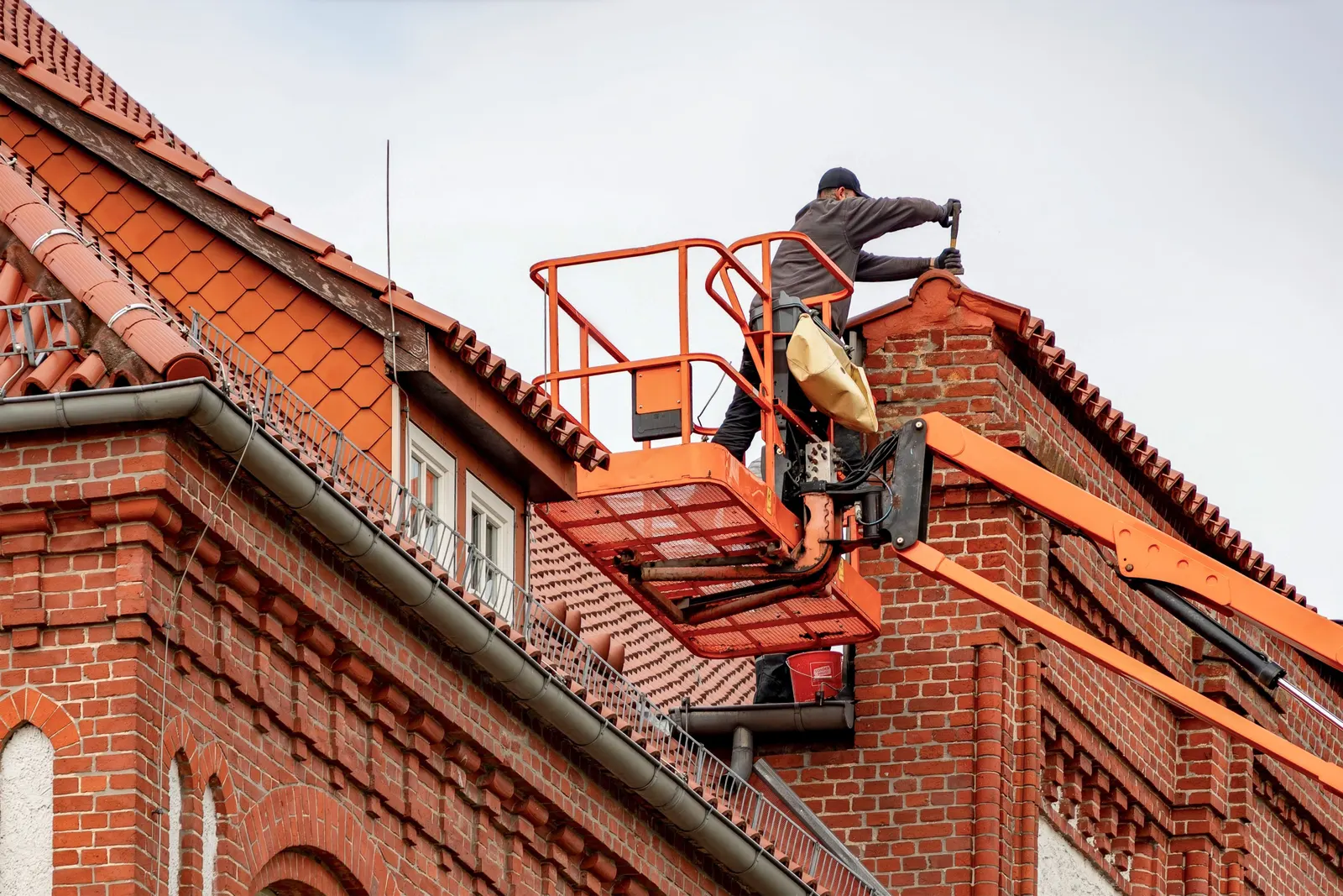 A worker on a raised platform working at the eaves of a tiled roof on a brick building.