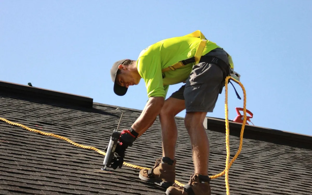 A roofer in a yellow shirt on a shingle roof.
