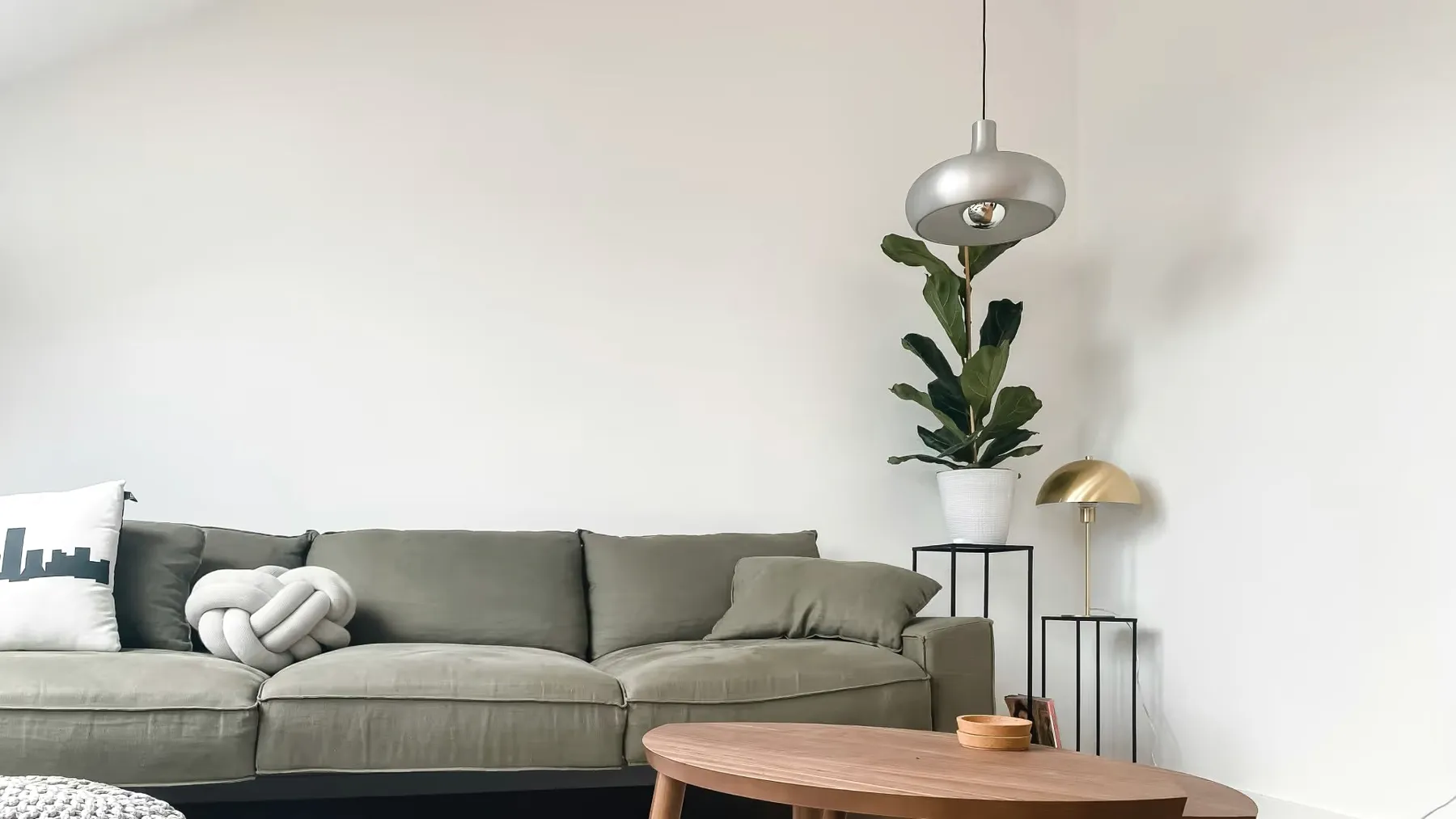 A calm living room: a green sofa, a round wooden table, a tall plant and a pendant lamp.