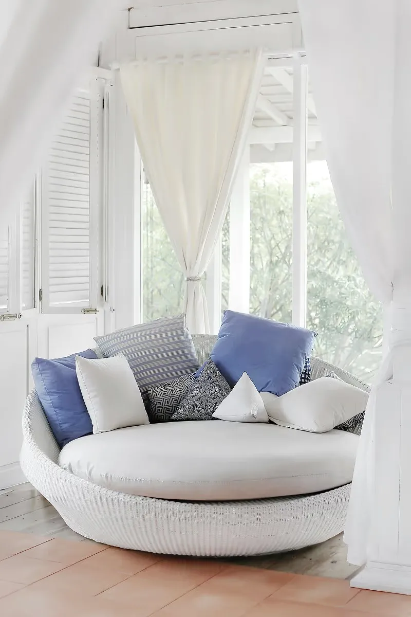 A round white wicker daybed with blue and white cushions by a curtained window.