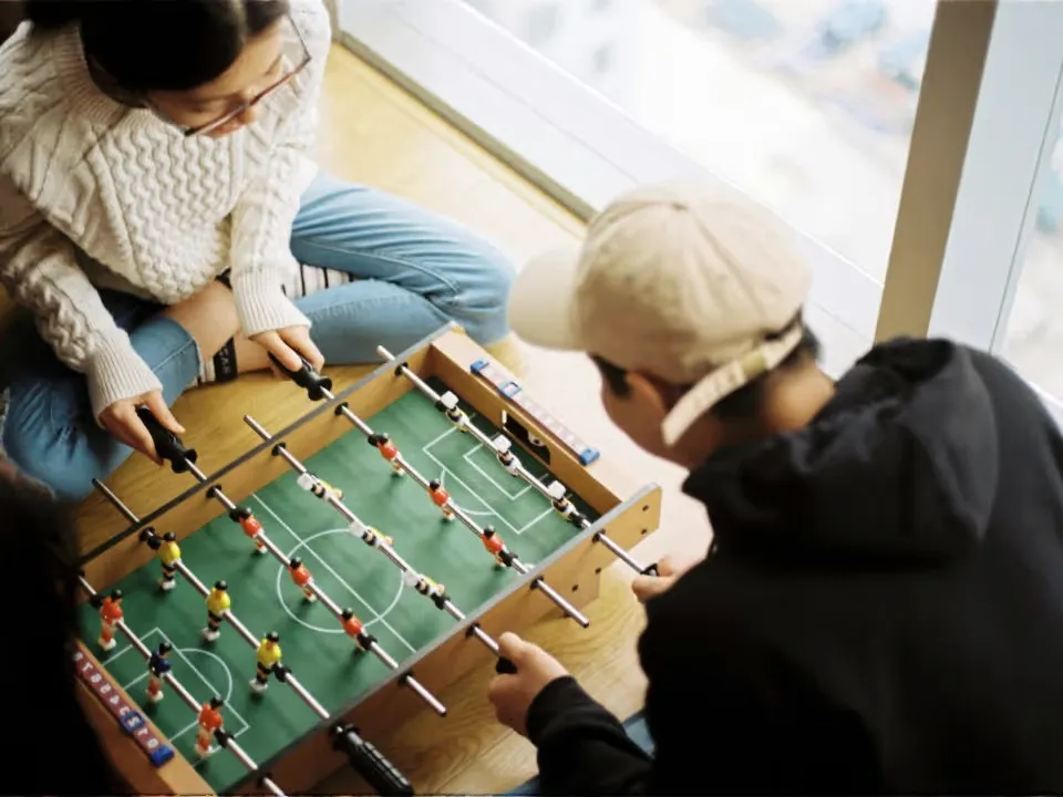 Two friends playing foosball by a window.
