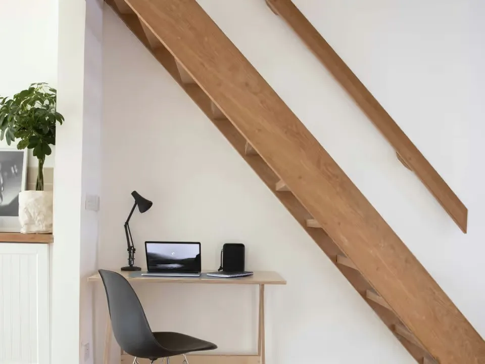 A small desk with a laptop under a wooden staircase.