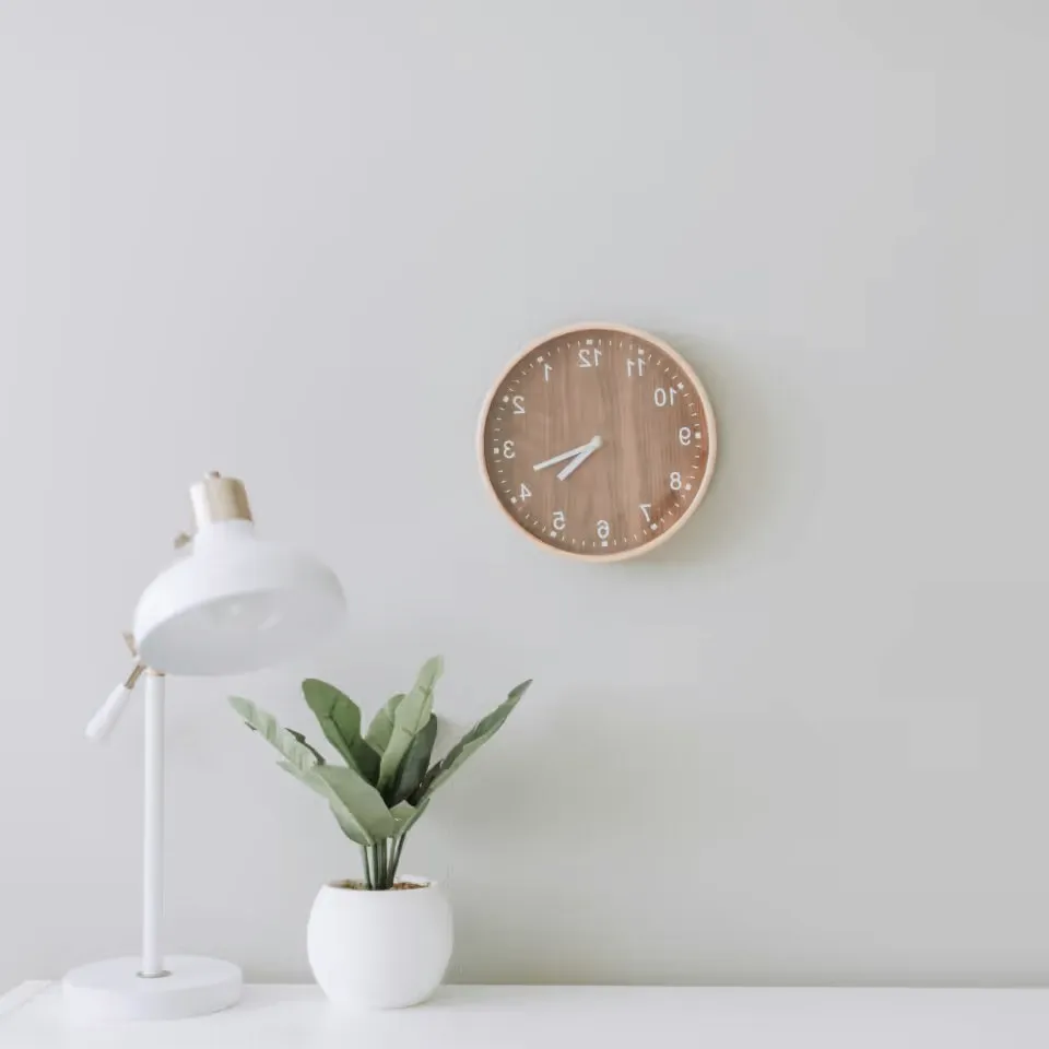 A round wooden clock over a white desk with a lamp and a plant.