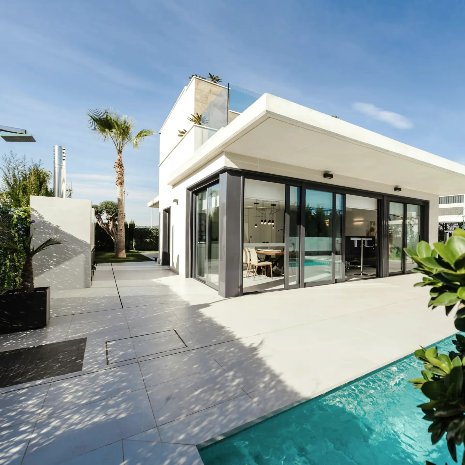 A white modern house with glass doors opening onto a stone terrace and a pool, a palm tree against a blue sky.