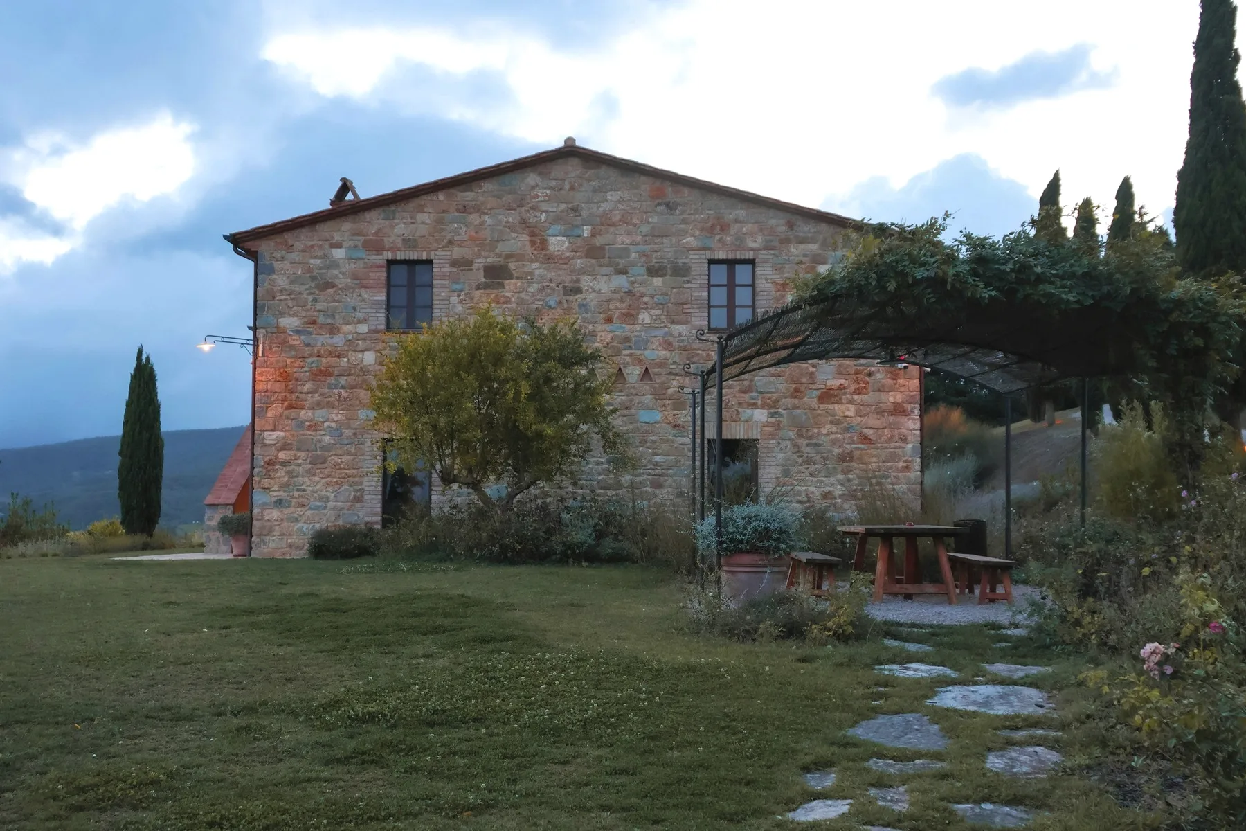 A stone villa at dusk, a pergola and a table on the lawn.