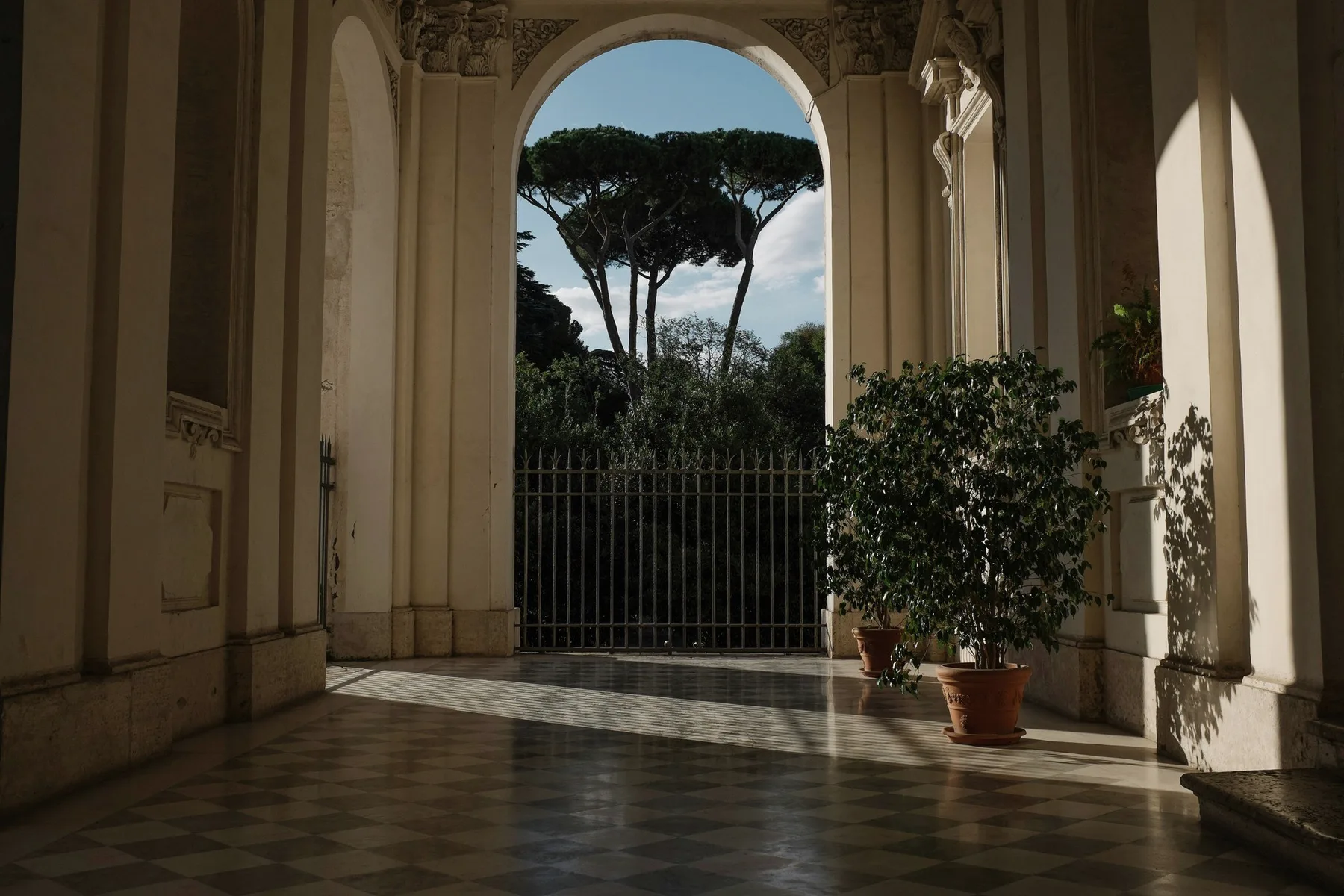 A stone arcade with potted trees, a gate and pines beyond.
