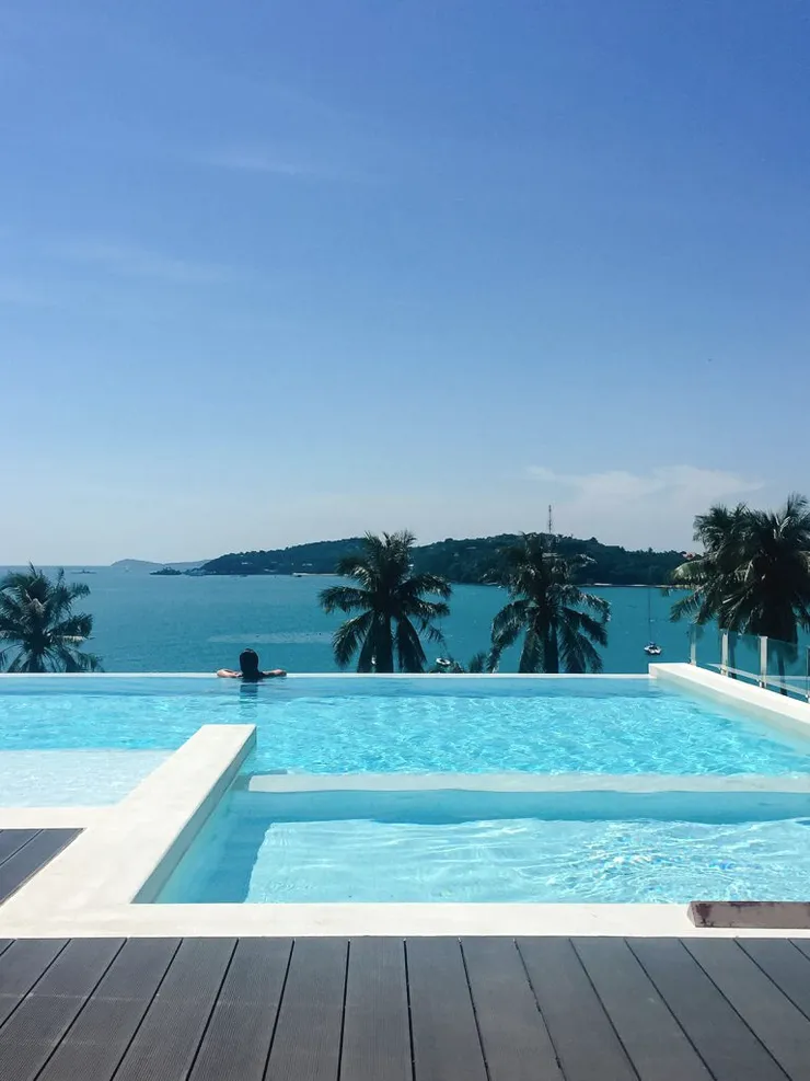 A pool on a wooden deck above a bay with palm trees.